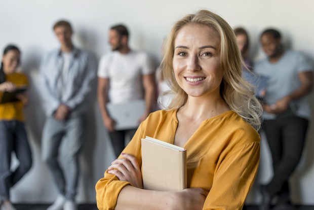 Blonde Frau mit Notizbuch lächelt in die Kamera, Kolleg:innen im Hintergrund.